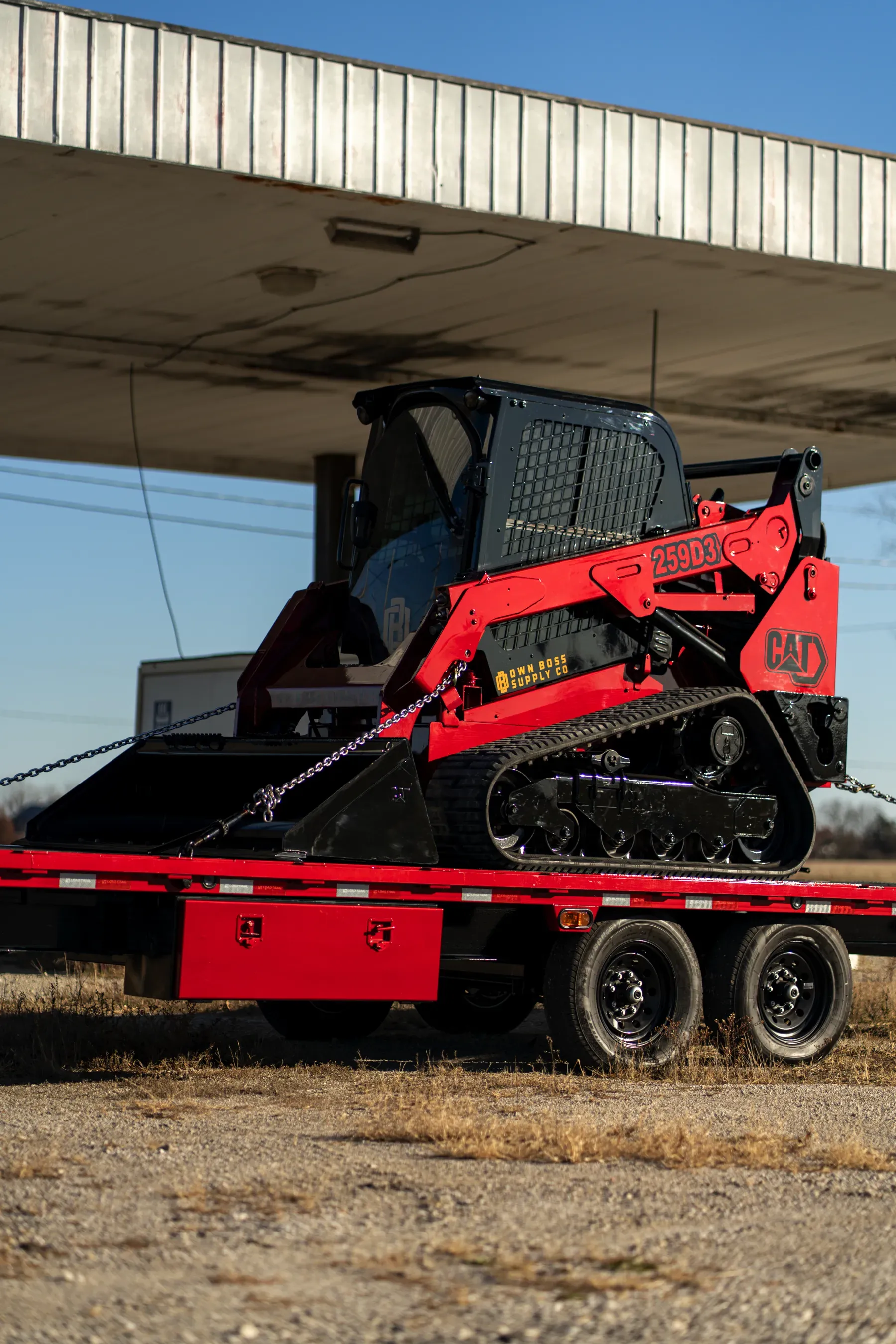 A red Bobcat compact track loader is on a flatbed trailer under a white overhang.