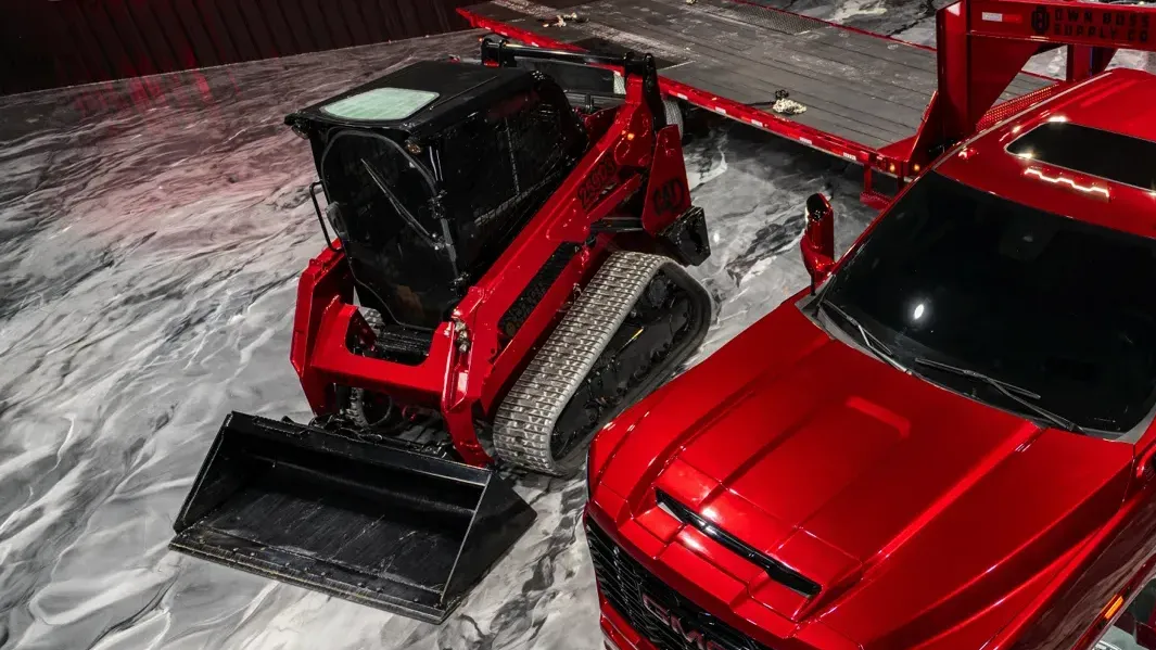 Shiny red pickup truck with a sunroof, parked next to a red compact excavator on a marbled floor.