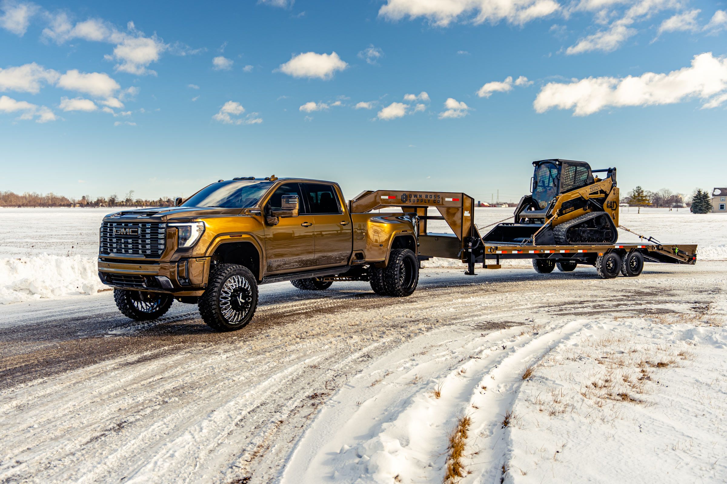 Bronze pickup truck towing a yellow skid-steer loader on a trailer through snow.