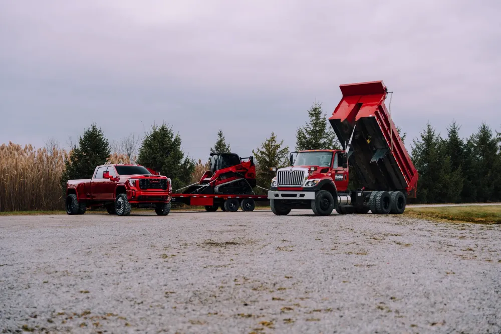 Red pickup truck, compact track loader on trailer, and dump truck on a gravel lot.