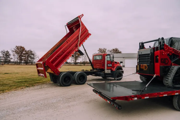 Red dump truck with lifted bed beside a red skid steer on a flatbed trailer.